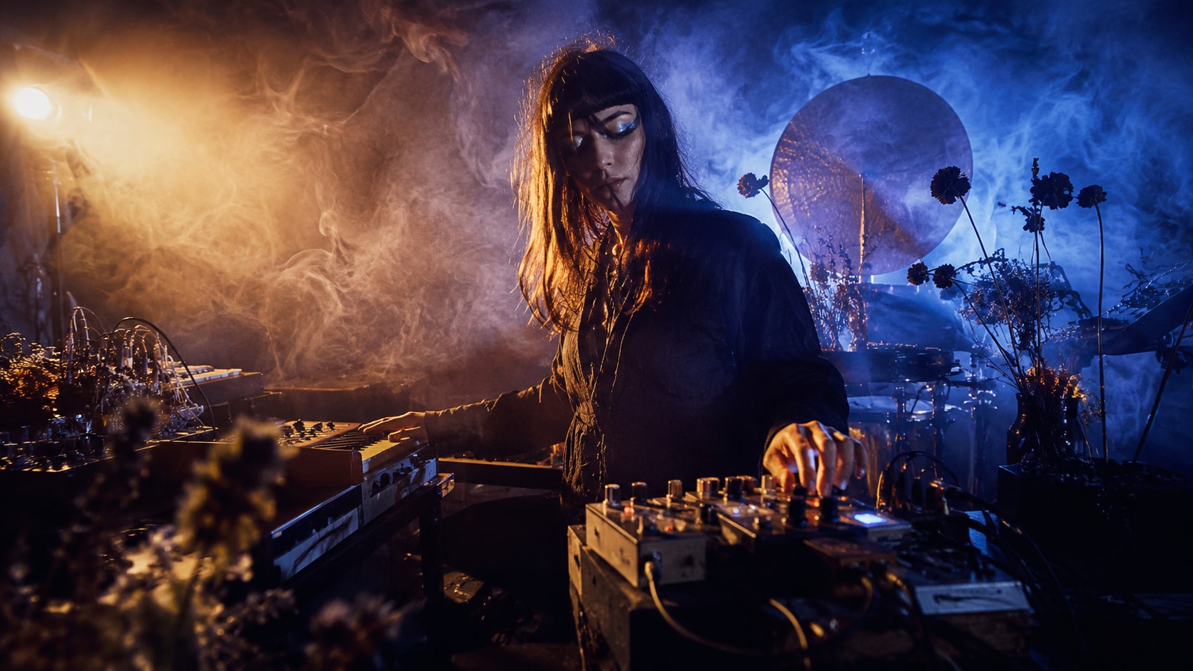 Carmen Villain adjusting electronic music gear in a dark, smoky studio with blue and warm lights.
