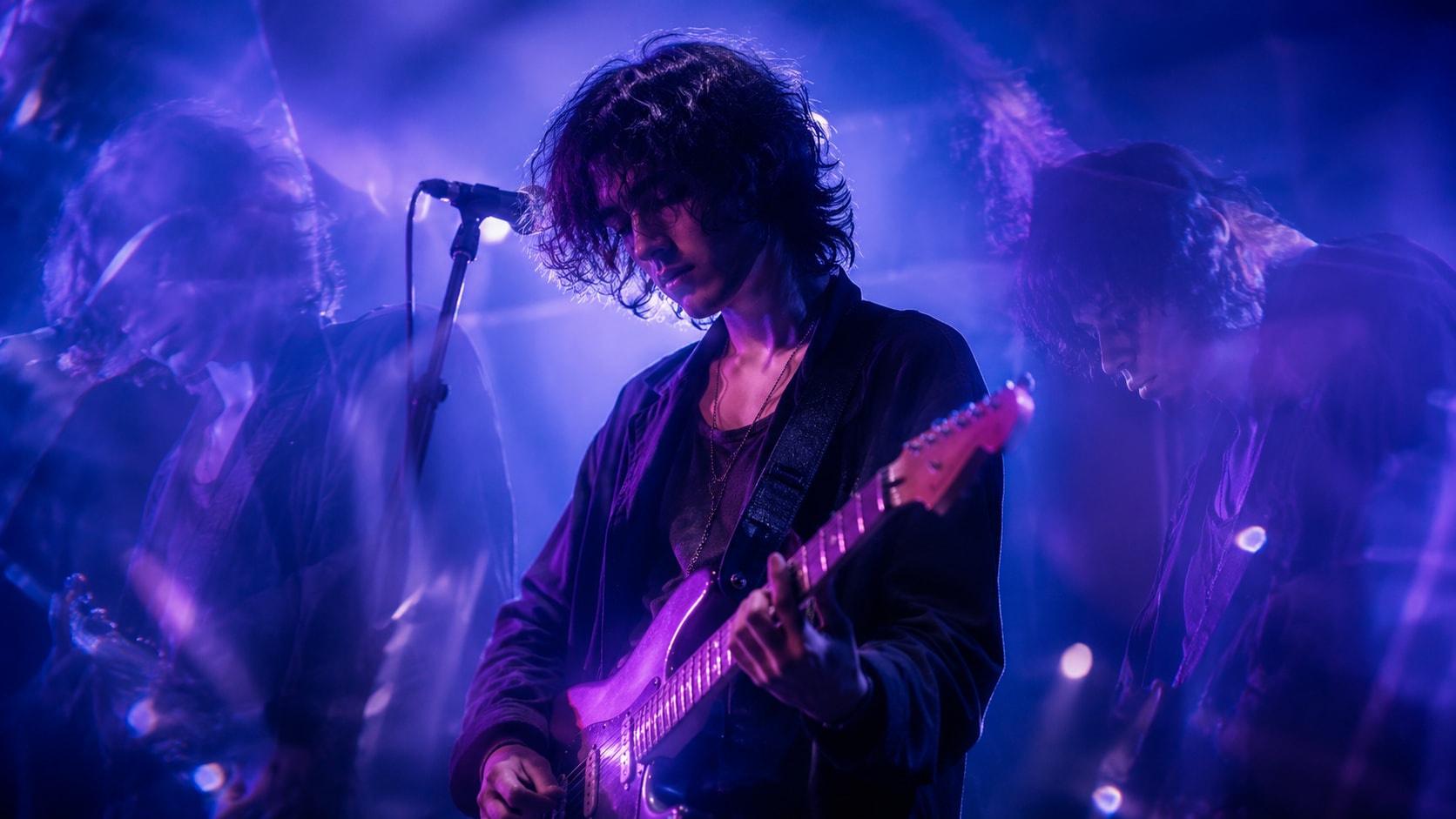 A young female indie musician with dark curly hair playing a purple electric guitar on stage under deep blue light.