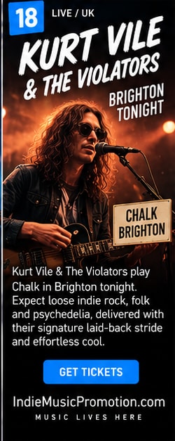 Kurt Vile playing electric guitar with shades on stage under moody lighting.